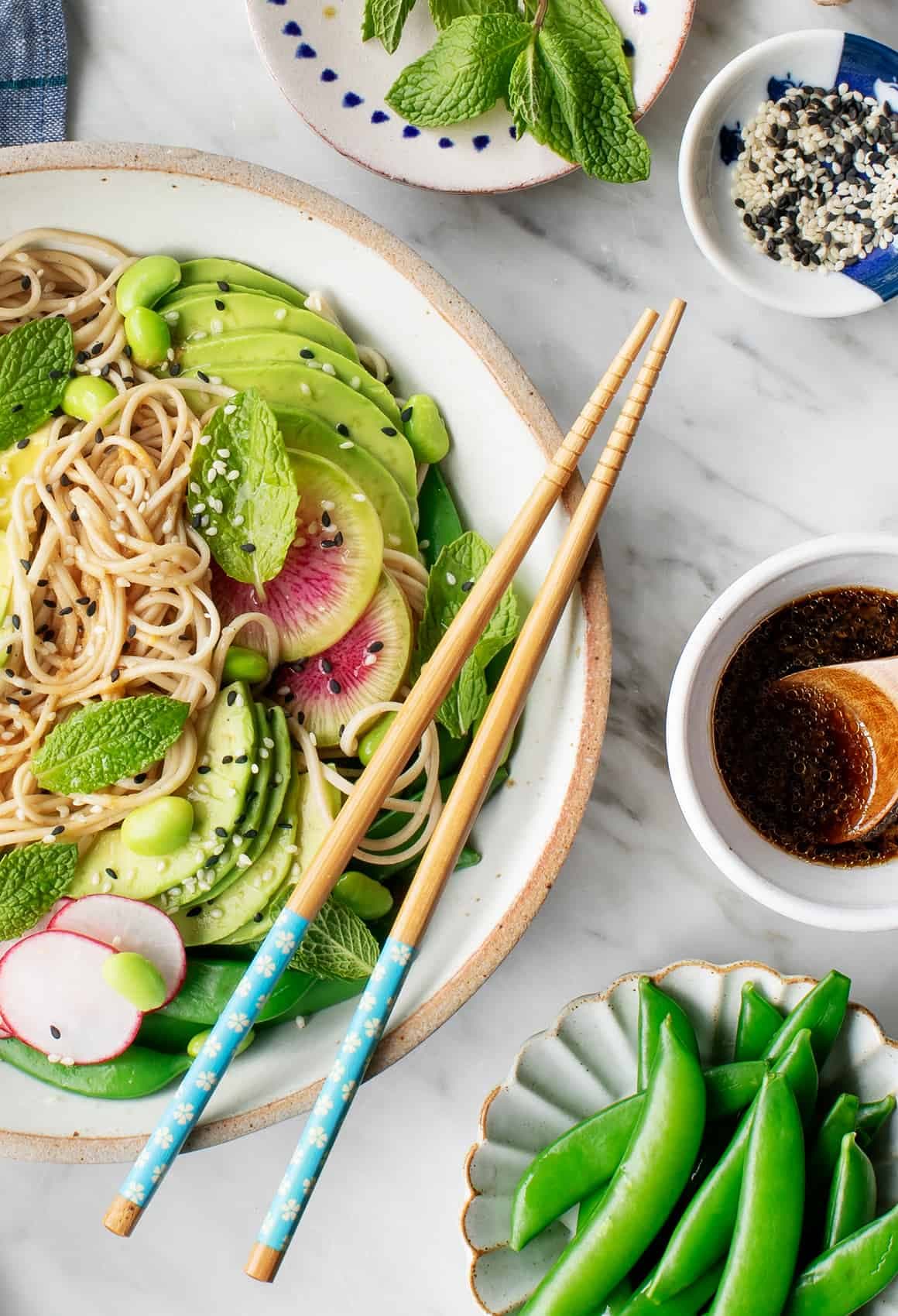 Noodle bowl next to sesame dressing, blanched snap peas, sesame seeds, and herbs