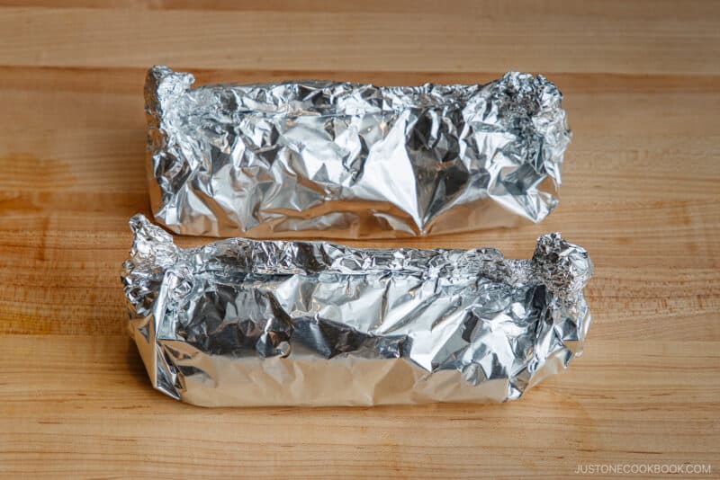 Two food items wrapped tightly in aluminum foil sit on a wooden surface. The foil is crinkled and shiny, completely covering the contents inside.