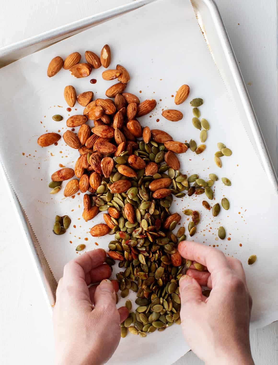 Hands tossing almonds and pepitas with seasonings on a baking sheet